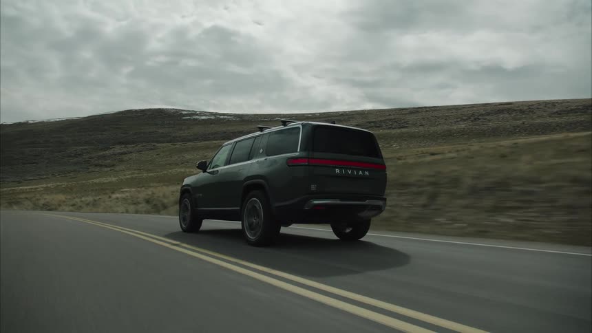 A darkcolored rivian r1s drives on an asphalt road through a barren landscape under a cloudy sky