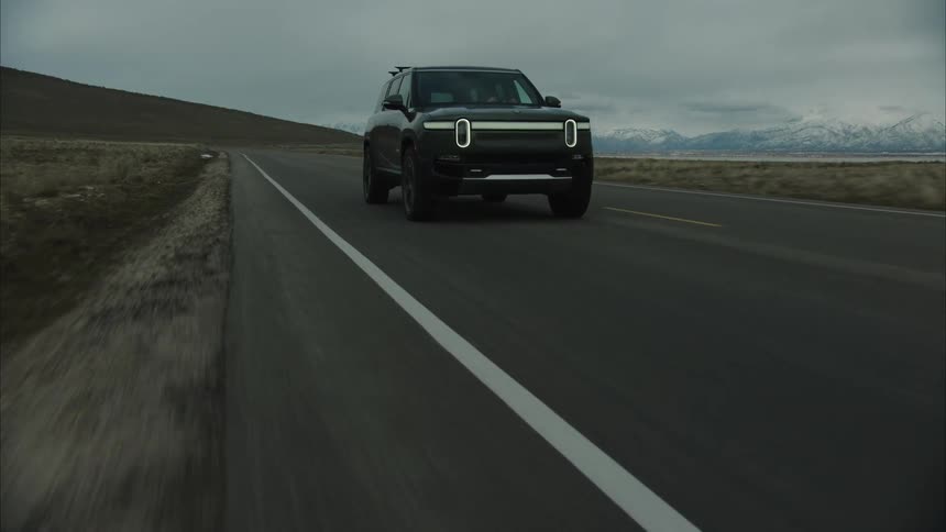 A dark rivian r1t drives along a straight asphalt road with a lake and snow capped mountains visible in the distance under a cloudy sky