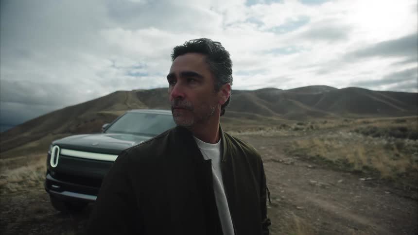 A man stands beside a parked car on a windy day surrounded by a countryside landscape with mountains under a cloudy sky