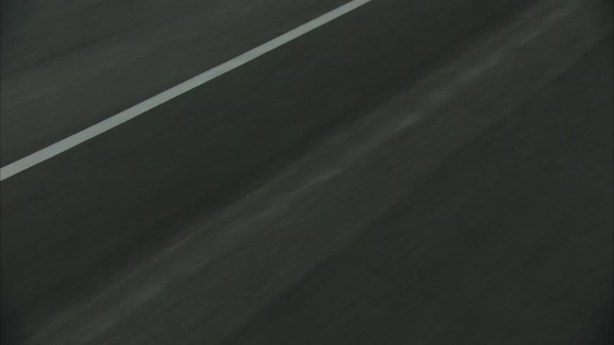 A black vehicle drives along a lakeside road passing a rest stop with parked cars set against mountains on an overcast day