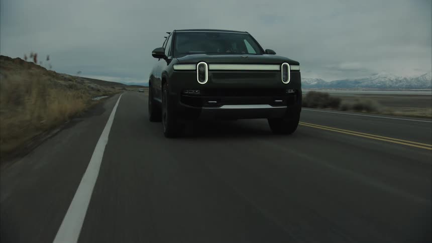 A darkcolored rivian r1s suv drives along a winding asphalt road through a barren landscape towards a lake with snowcapped mountains in the distance under a cloudy sky