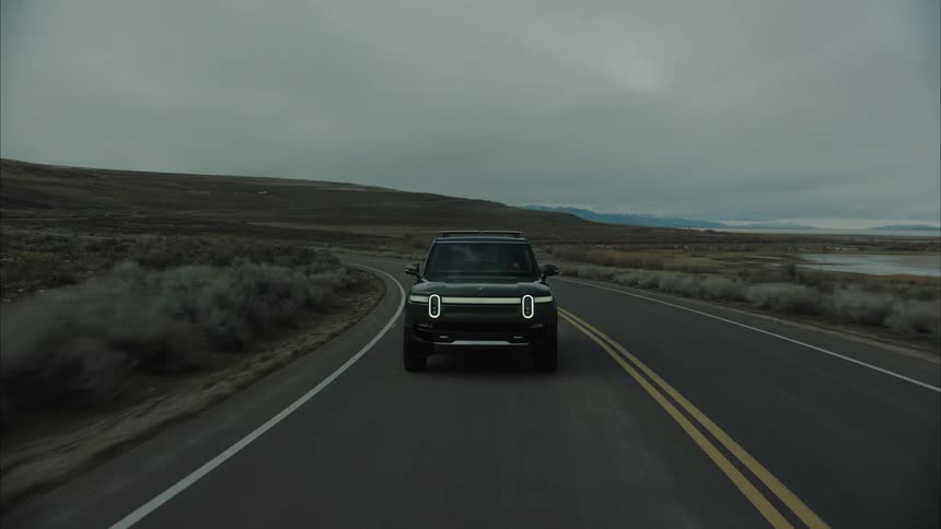 A rivian sport utility vehicle drives along a remote highway in a high desert on a cloudy day