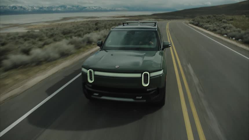 A dark rivian r1t drives along a straight asphalt road with a lake and snow capped mountains visible in the distance under a cloudy sky