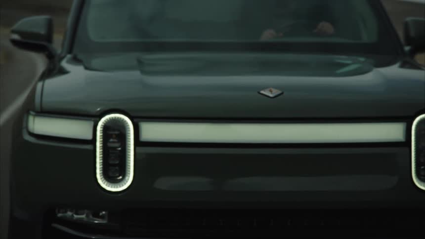 A dark rivian r1t truck shows its distinct front headlights and illuminated light bar
