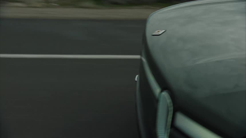 A dark green rivian suv travels on a paved road flanked by a lake heading toward a distant mountain range