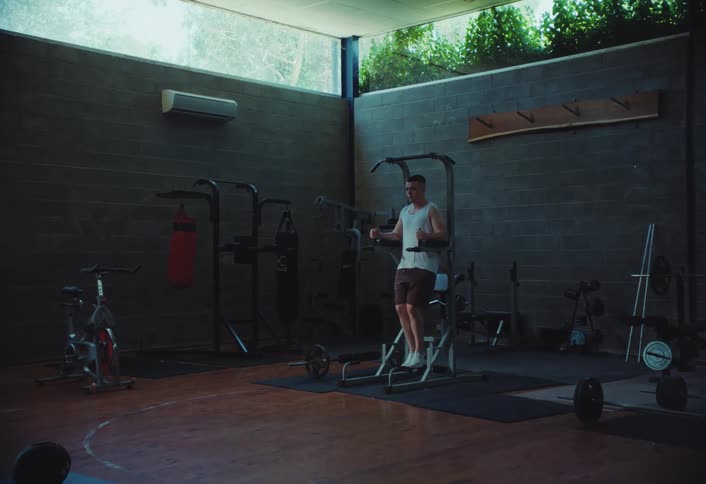 A young man performs leg raises on a dip station in a gym filled with various exercise equipment