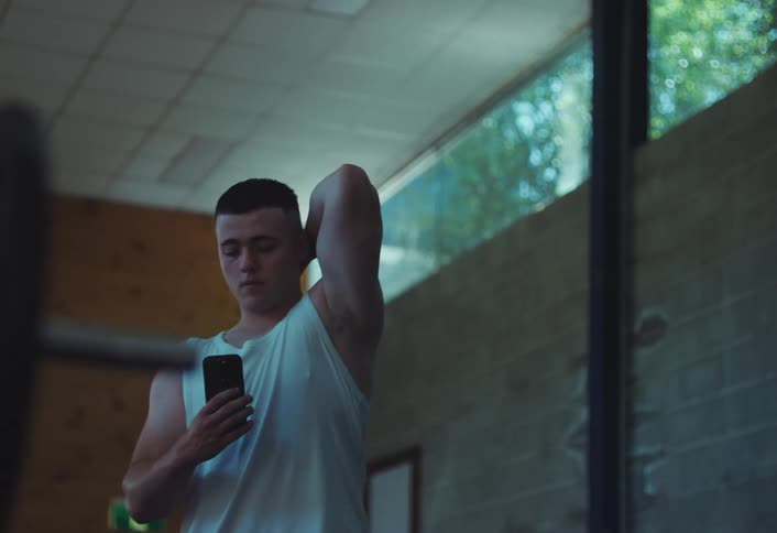 A man is posing in the mirror and texting on the phone while taking a break from weight training at the gym