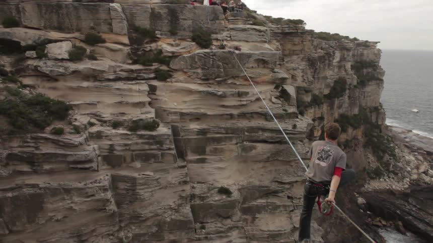 A man sits on a tight rope between mountains above the ocean