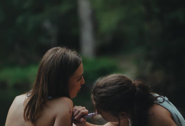 Two teen friends are laughing and drawing on each others shoulders with a pen while sitting on a riverbank