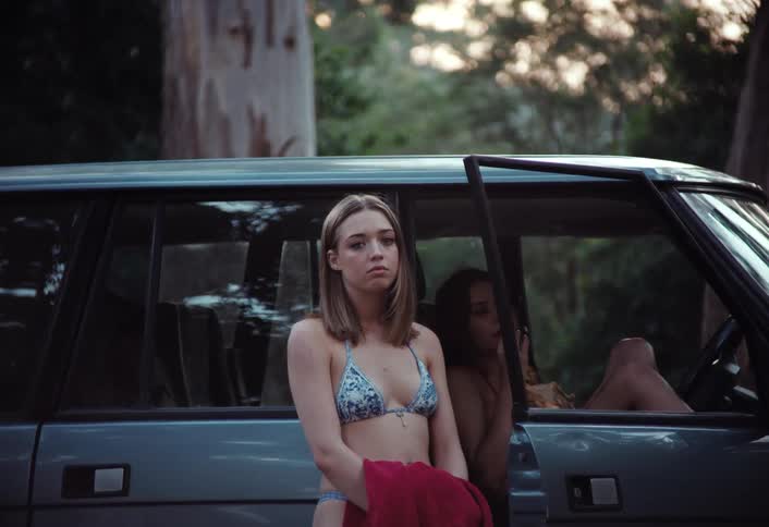 Two women one in a bikini and holding a towel stand next to a car with one of them sitting on the drivers seat
