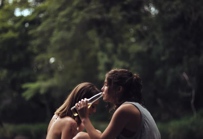 Girls in a park one drinks a beer and puts the mark with a pen the other eats a candy