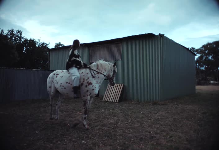 A woman rides a white horse with brown spots near a shack with metal walls set in a rural countryside