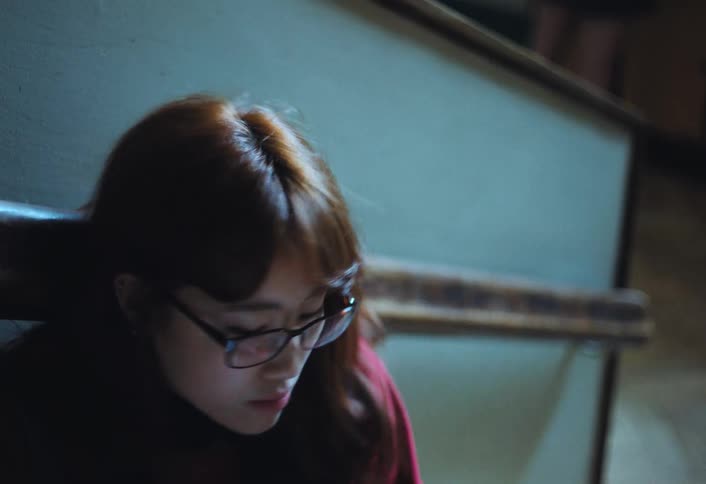 An asian girl wearing eyeglasses is scrolling on her phone while sitting on the stairs inside a school