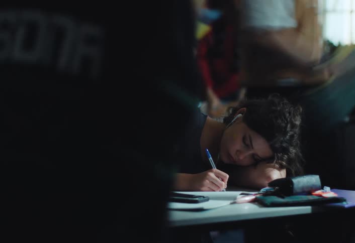 A teenage girl wearing headphones reclines at a desk in a classroom writing in a notebook as other students rush out