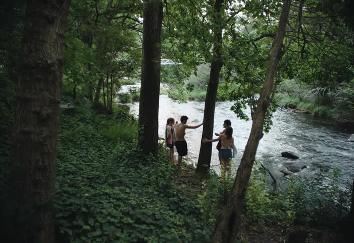 A group of friends stand near the edge of a flowing river surrounded by trees and ground cover