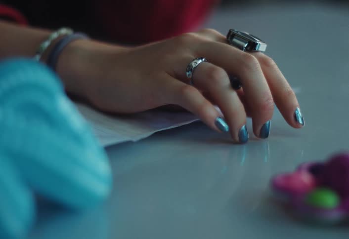 A closeup shows a hand with blue metallic fingernails tapping on a table adorned with rings and bracelets and holding a pen