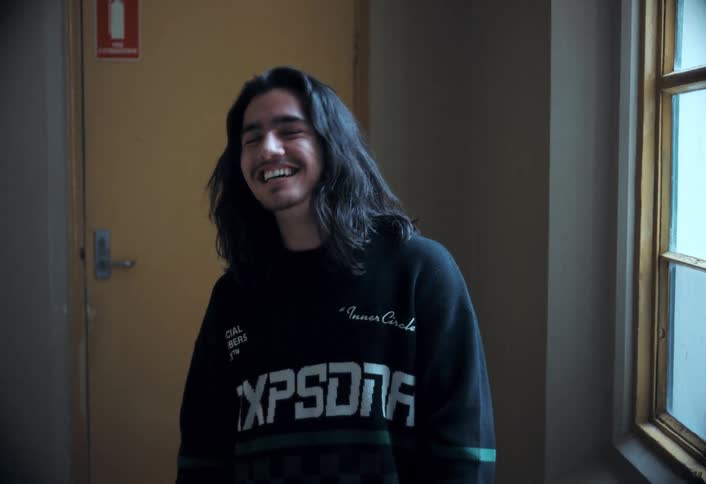 A young man with long hair smiles and laughs while standing in a room near a window