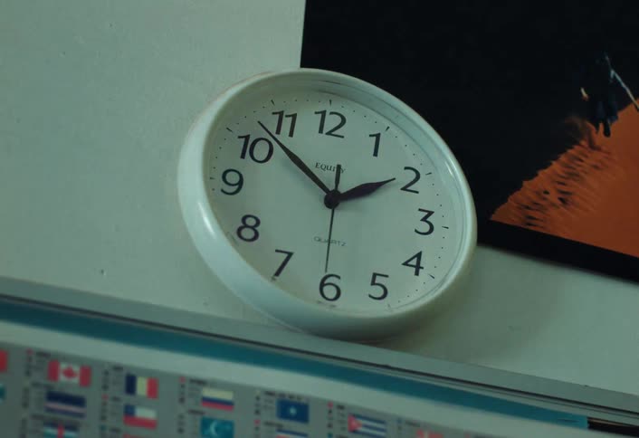 A round white clock with black numbers and hands is mounted on a lightcolored wall slightly angled with a blurry image of flags below it