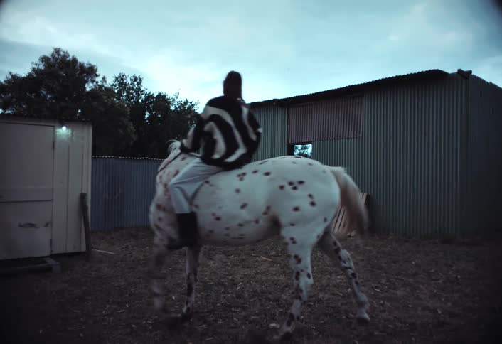 A woman rides a white horse with brown spots near a shack with metal walls set in a rural countryside