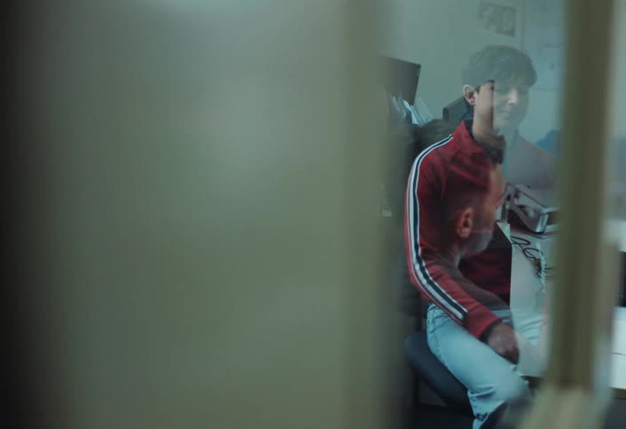 A boy in a red jacket sits in an office during an interview or meeting talking to an adult whose face is reflected on a window