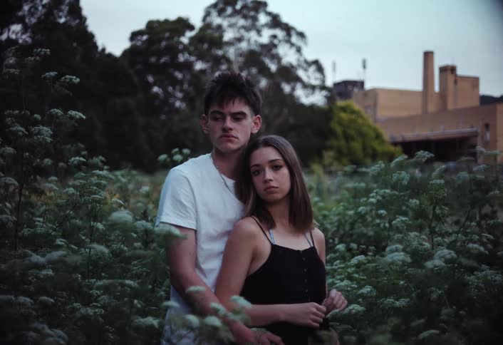 A young couple embraces as the man hugs the woman from behind in a garden filled with wildflowers swaying gently in the morning wind