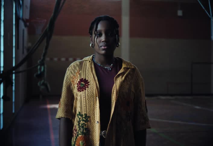 A young black woman stands confidently in a dimly lit gymnasium wearing a patterned shirt