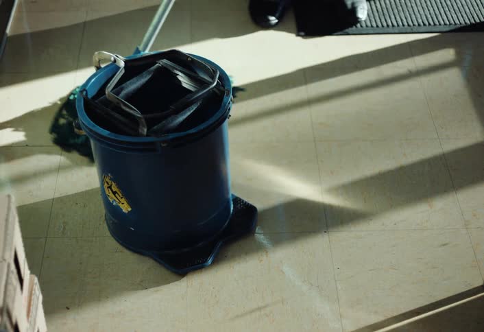 In a bright interior a person mops the tile floor while a blue bucket rests nearby and long shadows stretch across the surface as sunlight filters through