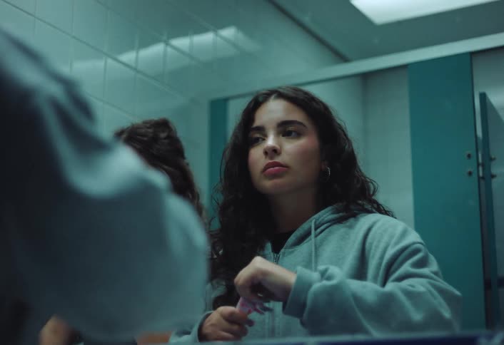 A young woman applies makeup in a public restroom checking her reflection while another person is seen beside her in the mirror