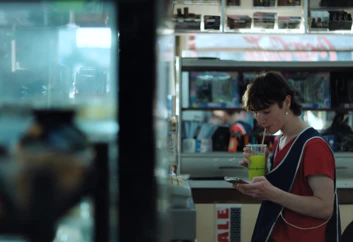 A store clerk takes a break browsing their phone while sipping a green frozen slushy beverage