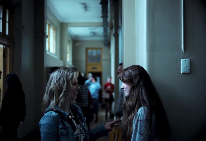 Two young women talk and smile in a school hallway while other people move in the background
