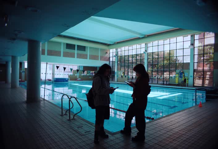 Two friends stand by an indoor pool engrossed in their phones as sunlight streams through large windows in the background