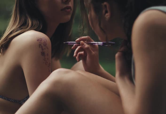 Two girls sit on the grass one is drawing a tattoo on the others shoulder with a pen while her wet hair shows shes been swimming