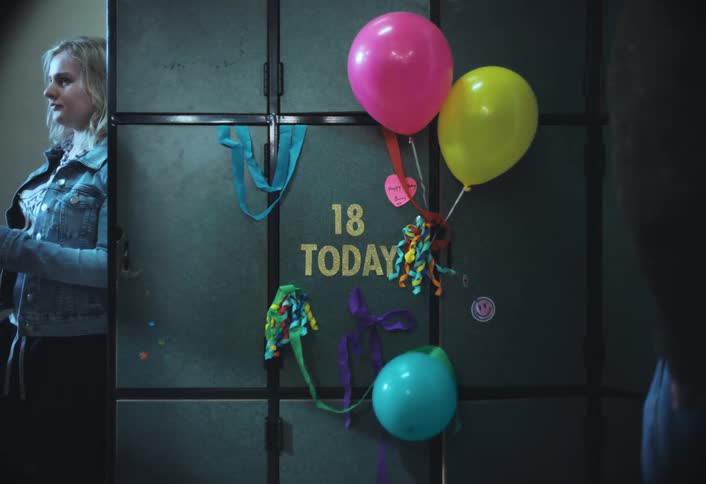 A pink yellow and pale blue balloon float in front of a wall that has the words 18 today in yellow while a young blonde woman in a jean jacket speaks to a person out of view as other people mingle and walk past the camera