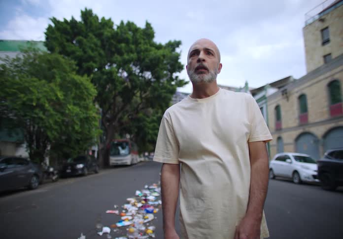 A frustrated person stands on a littered street holding two large black garbage bags before angrily tossing them onto the ground