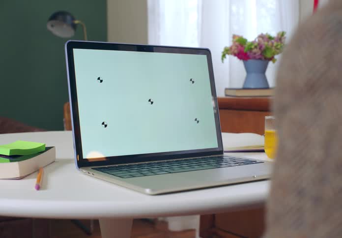 A laptop with a replaceable white screen rests on a table in a brightly lit home office while a person gestures in front of it