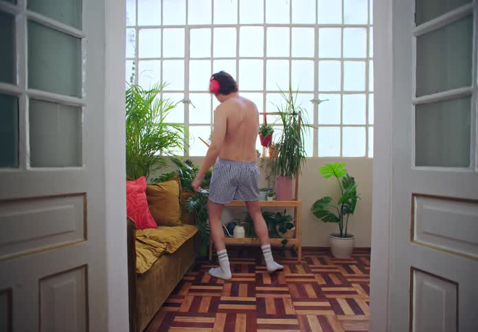A woman working on a laptop turns and looks irritated at a man dancing in his underwear while watering plants