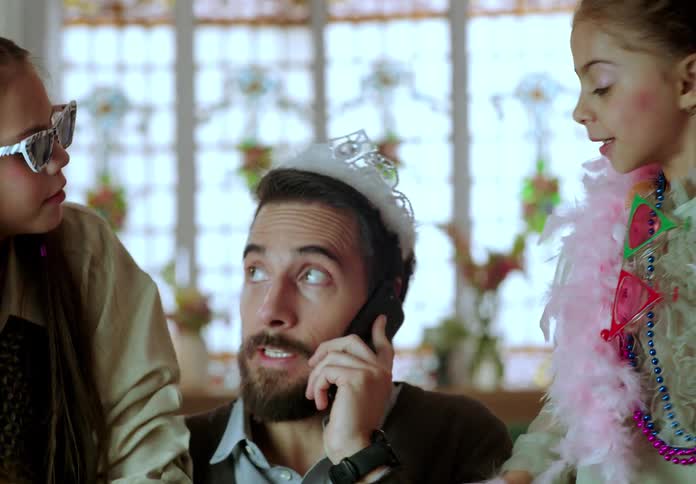 A father attempts to take a call on his smartphone while his daughters playfully dress him up with a tiara and makeup
