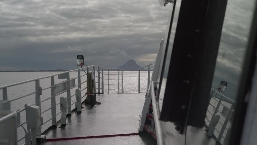 From a vantage on the deck the boat moves forward across calm waters under an overcast sky with the silhouette of a distant mountain visible on the horizon