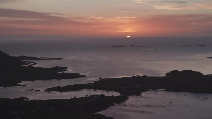From a lofty vantage the sun dips toward the horizon in hues of orange and pink illuminating the winding coastline and small islands below while scattered houses and tranquil waters complete the serene evening panorama