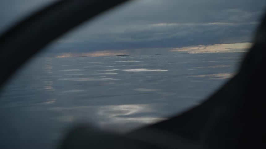 A view of the ocean horizon and cloudy sky is framed through a dark curved window of a vehicle
