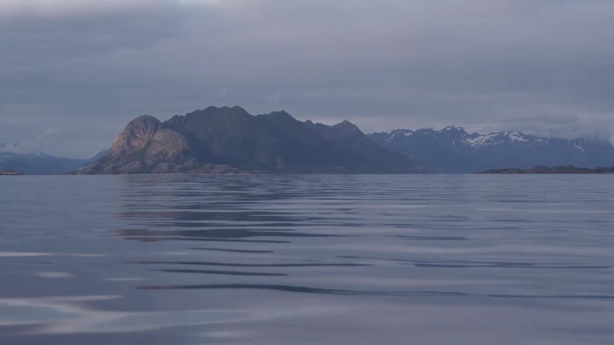 Smooth water ripples softly under a subdued sky reflecting the outline of distant mountains as gentle waves create a subtle sense of motion across the serene surface