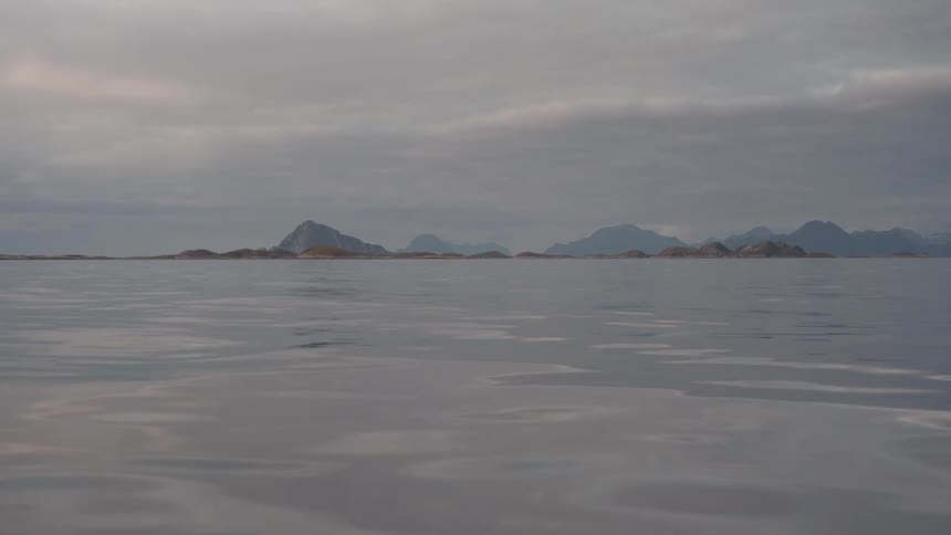 A tranquil and expansive seascape with calm open waters beneath an overcast hazy sky reveals a distant chain of small soft rocky isles along the faroff skyline with gentle ripples on water