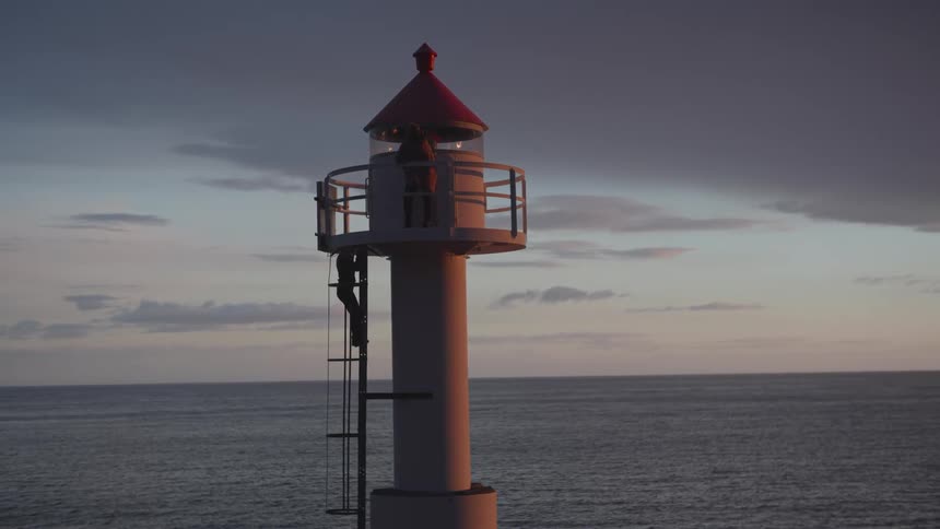 A couple standing over a lighthouse she climbs the stairs at dusk or dawn with a little wind