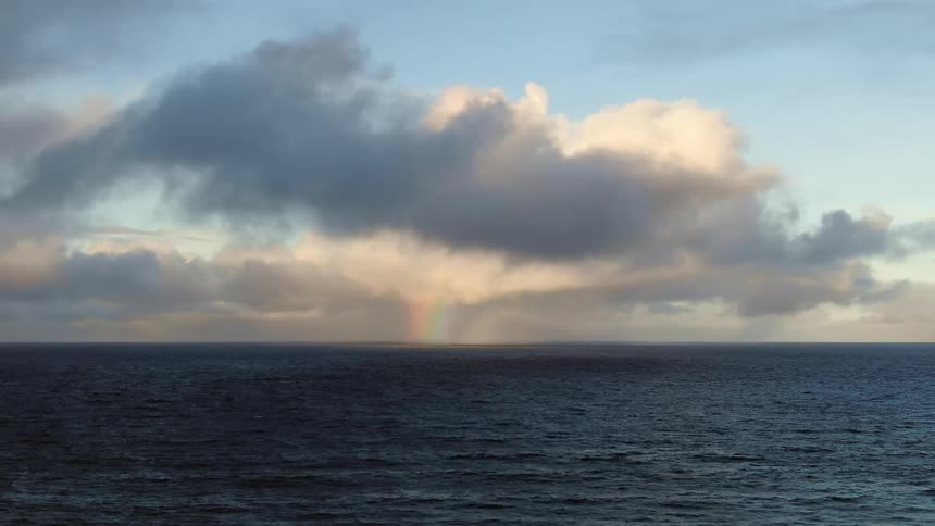 A distant rainbow appears in a gap between storm clouds over a dark choppy sea