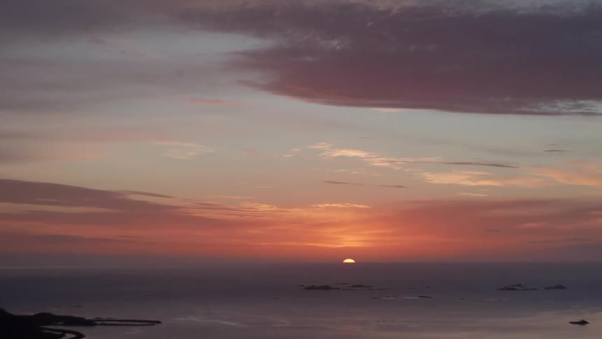 From a lofty vantage the sun dips below the horizon in warm hues of orange and pink casting a gentle glow across small islands and serene waters while softly lit clouds linger above