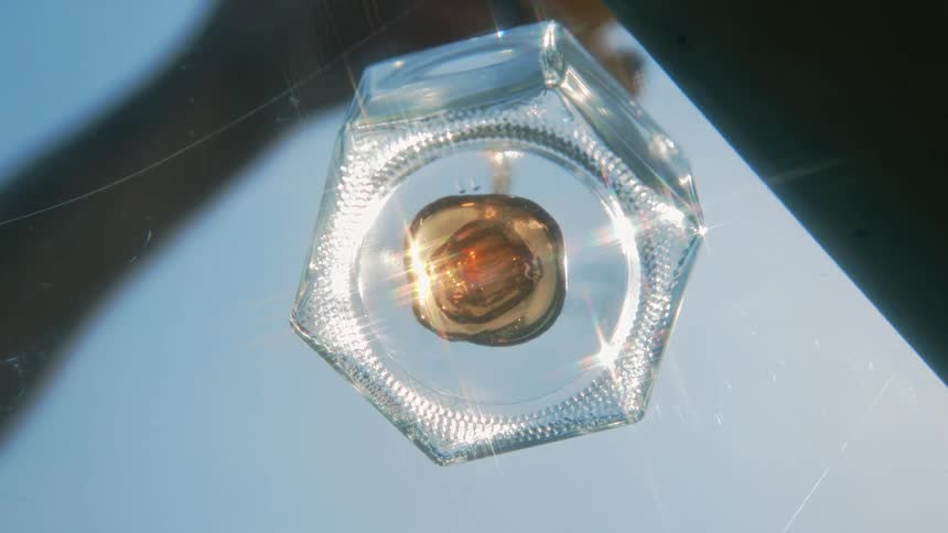 Honey pours inside a clear hexagonal glass bottle reflecting light under a clear blue sky