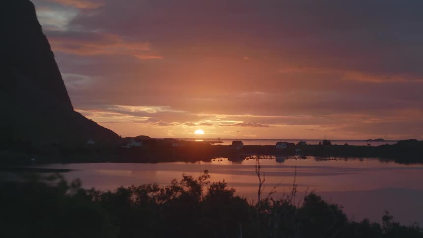 The sun sets over a calm body of water reflecting a vibrant orange and purple sky with a village nestled along the shore and a mountain silhouetted on the left