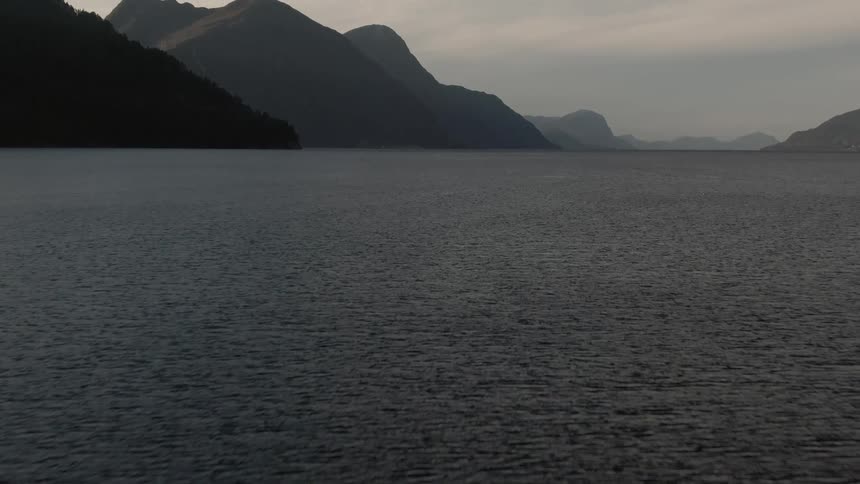 A vast dark body of water fills the frame stretching towards silhouetted mountains in the distance under an overcast sky