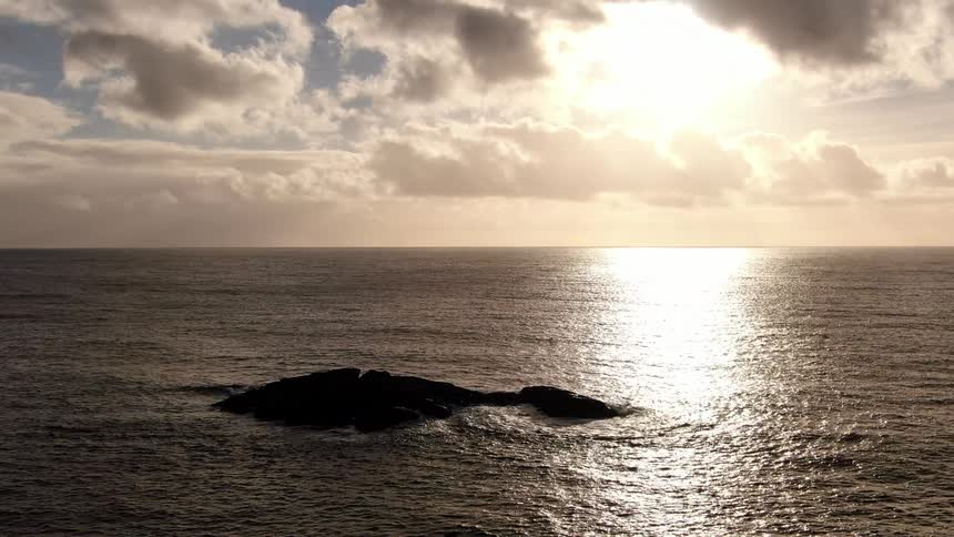From an elevated perspective the camera moves toward a small rocky island at sunset with golden light reflecting on the oceans surface and scattered clouds drifting across the sky in a tranquil coastal view