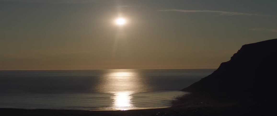 The sun shines brightly over a calm sea reflecting a shimmering path of light towards the viewer with a dark cliffside silhouetted in the foreground